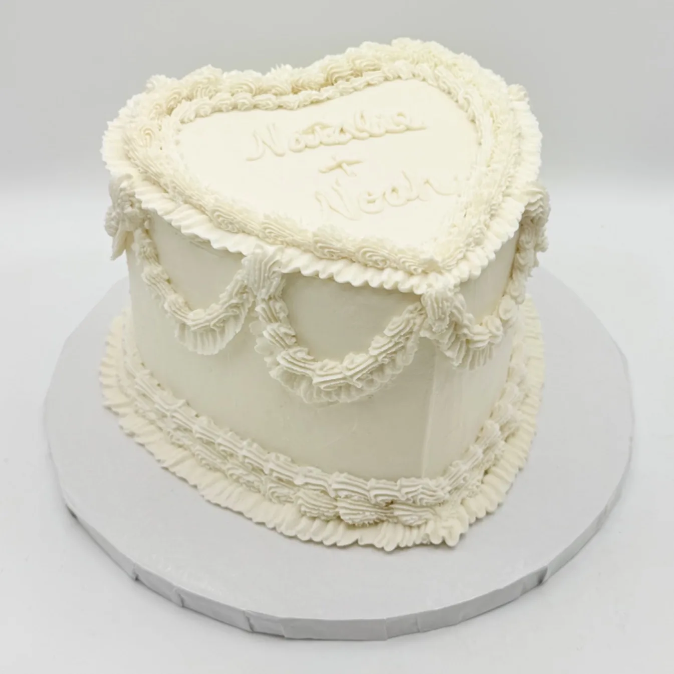 Natalie heart-shaped ivory buttercream wedding cake with piped shell borders and ruffled swag drapes around the sides, two names piped in script across the top, 2026