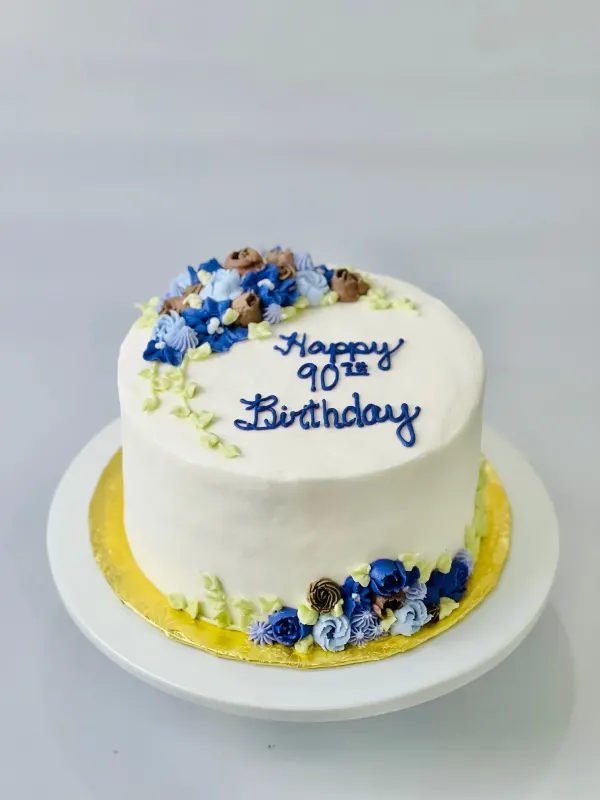 White cake with blue and mocha buttercream flowers and Happy 90th Birthday piped in navy