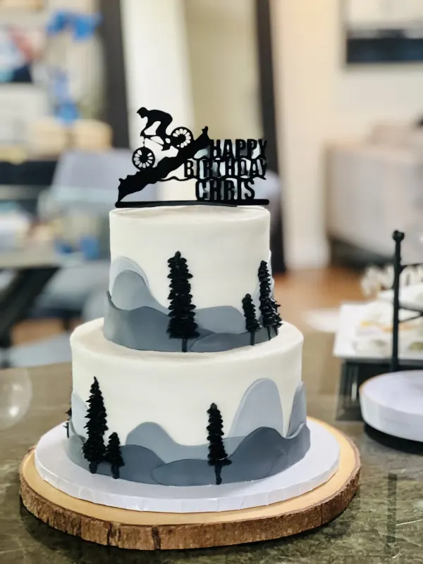 Two-tier mountain cake with grey peaks, pine trees and a mountain biker Happy Birthday Chris topper
