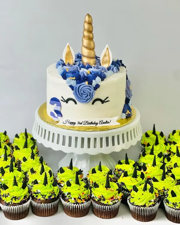 Unicorn cake with a gold horn and blue buttercream mane beside green unicorn cupcakes, Happy 3rd Birthday Analia