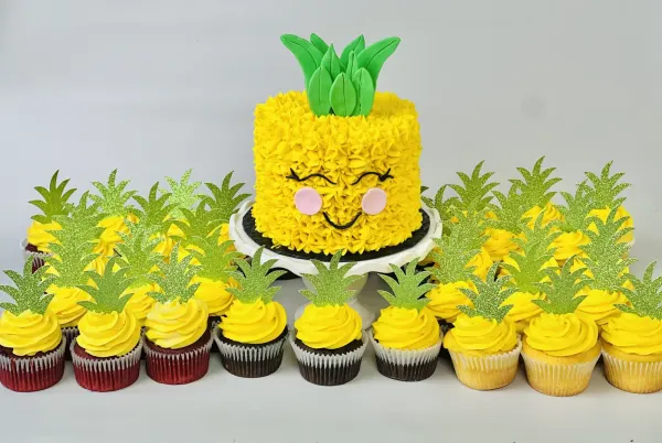 Pineapple birthday cake with piped yellow frosting, a smiling face and matching pineapple cupcakes