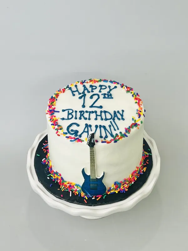 White cake with rainbow sprinkles, a blue electric guitar cutout and Happy 12th Birthday Gavin in blue
