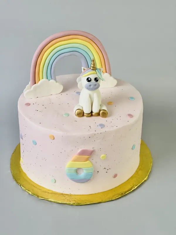 Pale pink unicorn cake with a pastel fondant rainbow and a seated unicorn with gold hooves