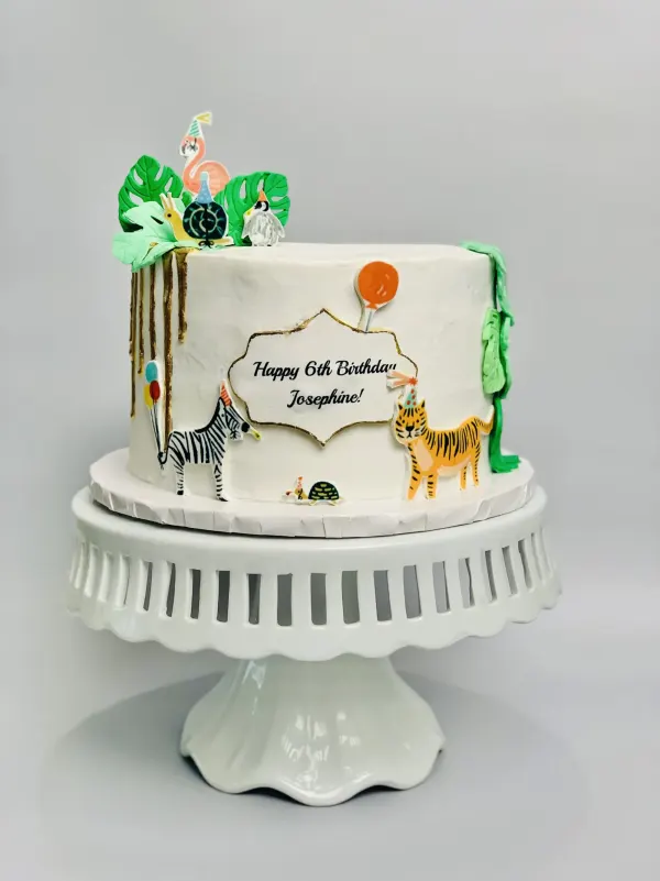 White safari cake with a gold drip, party-hat animal cutouts and Happy 6th Birthday Josephine