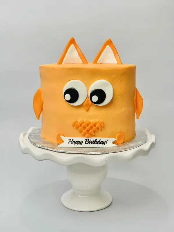 Orange owl-shaped birthday cake with fondant eyes, ears and feathers and a Happy Birthday banner
