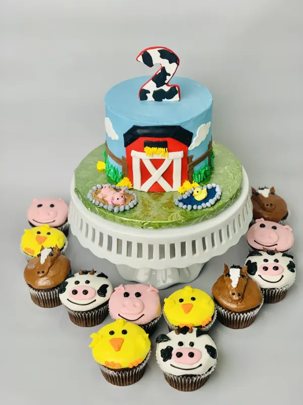 Barnyard second birthday cake with a red barn and cow-print 2, plus farm animal face cupcakes