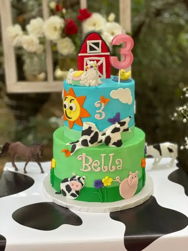 Two-tier farm birthday cake with a red barn, cow-print bow, pink 3 candle and the name Bella