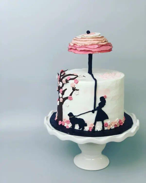 White cake with a navy silhouette of a girl walking a dog and a pink ruffled parasol topper