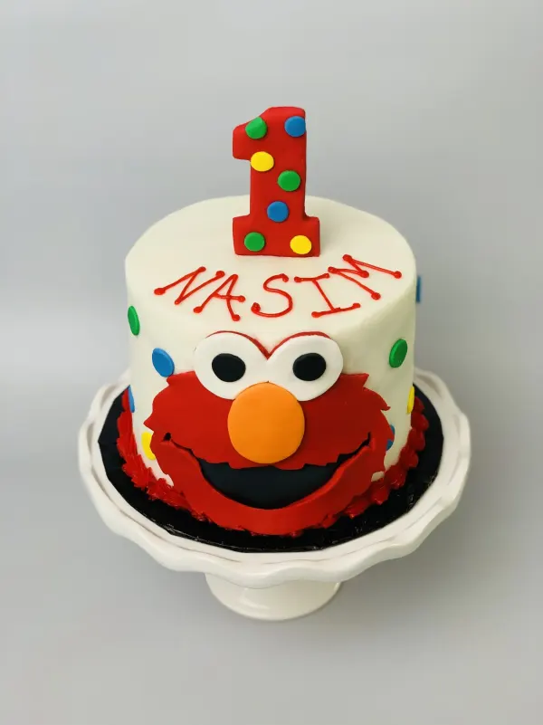 Elmo face birthday cake with a fondant number 1, colored dots and the name Nasim piped in red