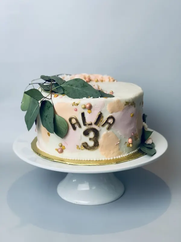White cake with blush abstract patches, gold leaf, eucalyptus leaves and gold Alia 3 letters