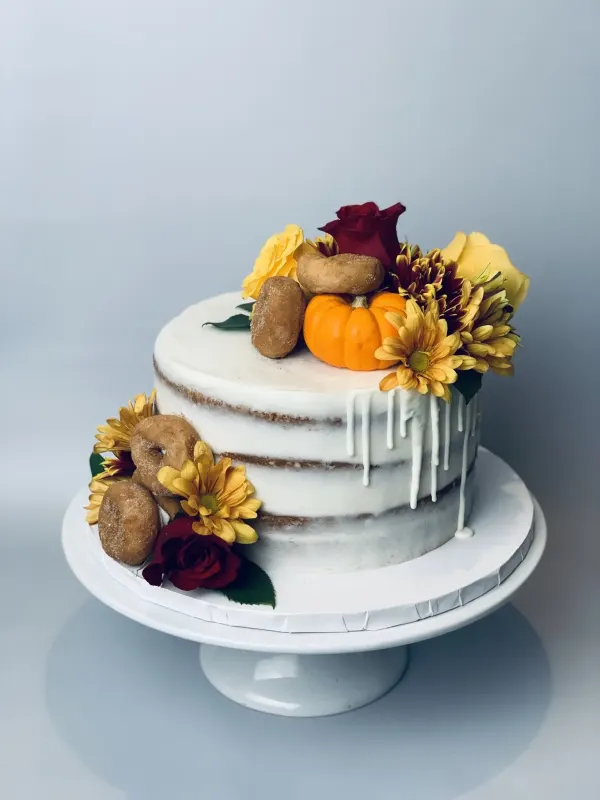 Semi-naked autumn cake with white drip, a mini pumpkin, cider doughnuts and yellow and red flowers
