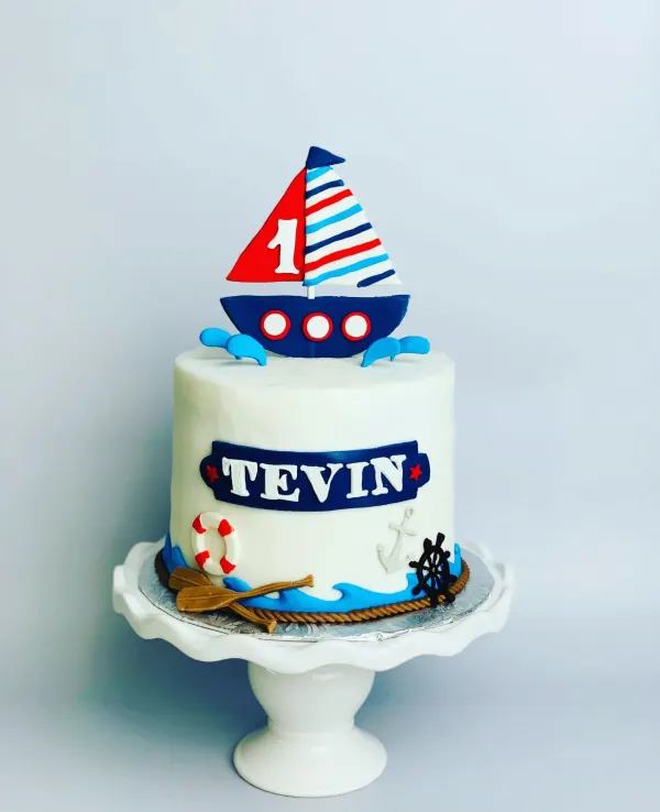 Nautical first birthday cake with a sailboat topper, anchor, life ring and a plaque reading Tevin