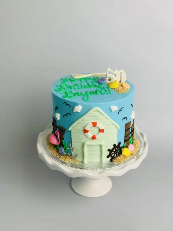 Beach hut 50th birthday cake with a life ring, flamingo, sand and Happy Birthday Bryan piped in green