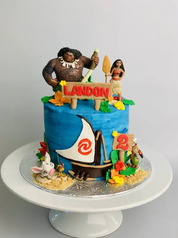 Moana birthday cake with Maui and Moana figures, a canoe and a wooden sign reading Landon