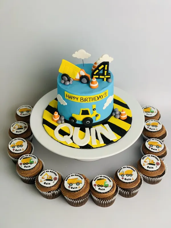 Construction birthday cake with a dump truck, digger and the name Quin, plus truck-topper cupcakes