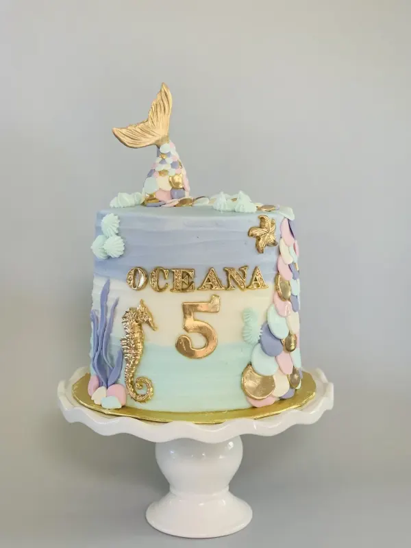 Mermaid birthday cake with a gold tail topper, seahorse and scales, reading Oceana 5 in gold