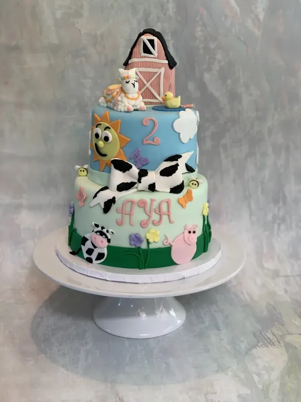 Two-tier farm birthday cake reading Aya with a barn topper, lamb, cow, pig and a number 2