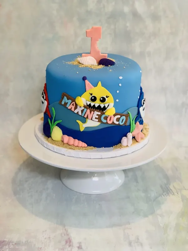 Baby shark first birthday cake with Maxine Coco on a driftwood sign, sand, shells and a pink number 1