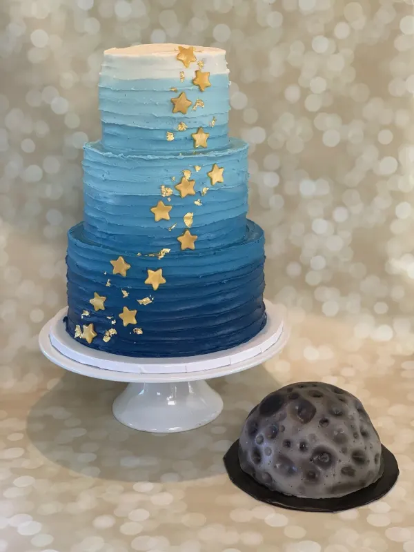 Three-tier blue ombre buttercream cake with gold stars beside a small grey moon cake