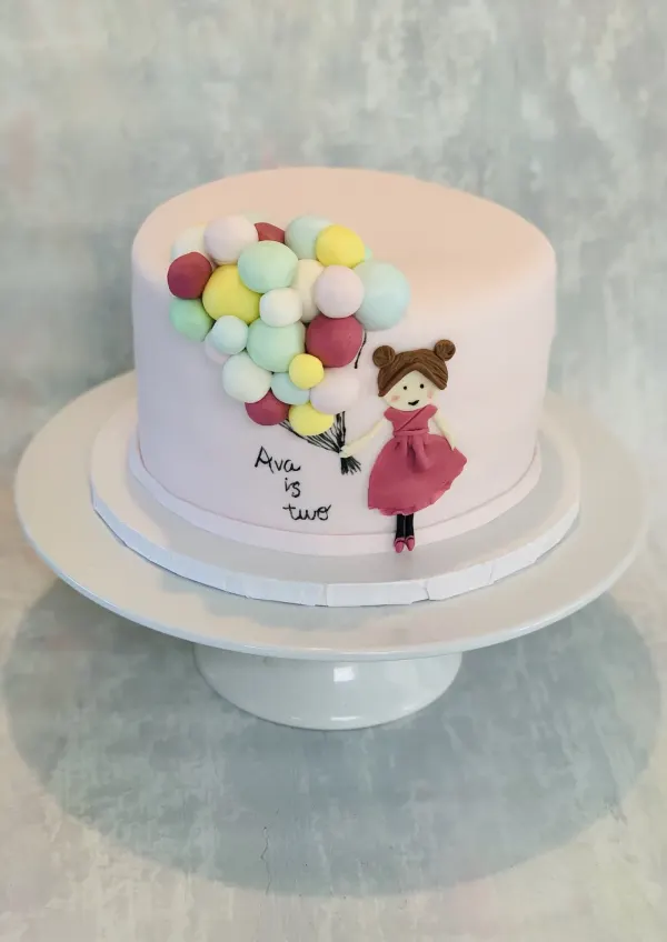 Pink birthday cake reading Ava is two with a fondant girl holding a cluster of pastel balloons