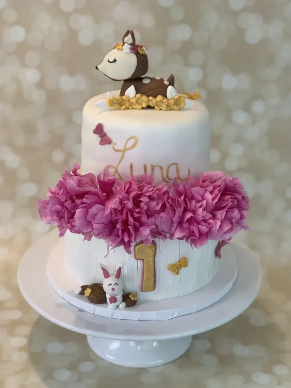 Two-tier white first birthday cake with a fawn topper, pink peonies, gold Luna script and a bunny