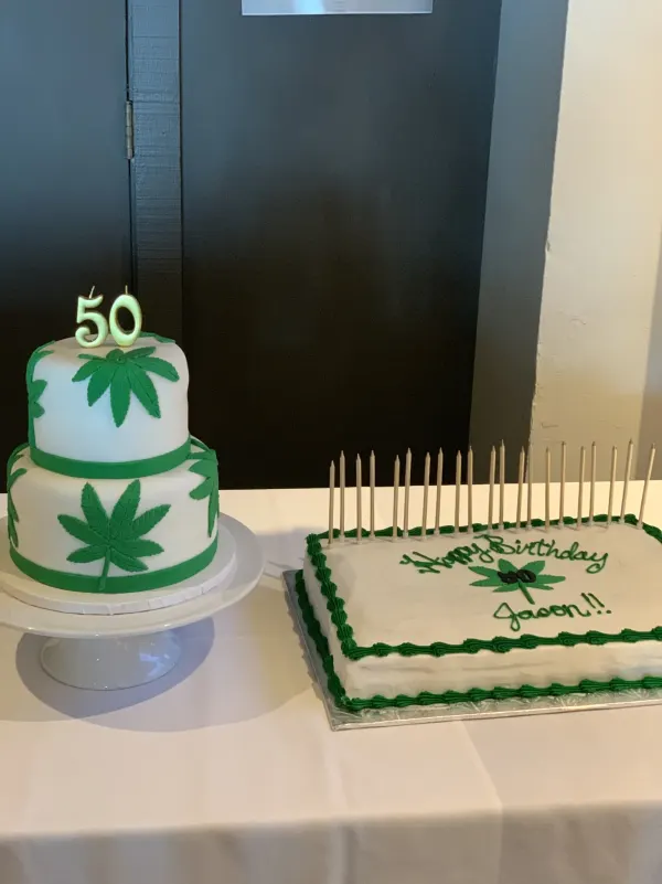 Two-tier white cake with green leaf decorations and 50 candles beside a Happy Birthday Jason sheet cake
