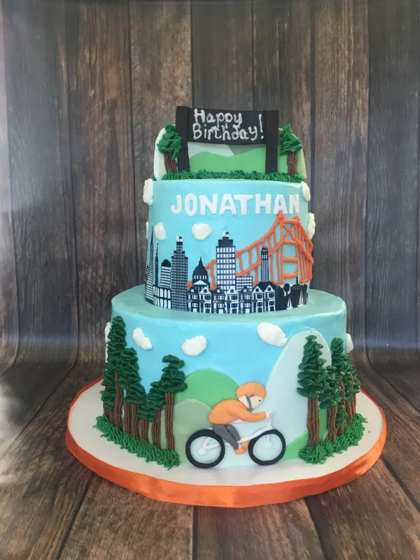 Two-tier buttercream cake with a San Francisco skyline, redwoods, a cyclist and piped JONATHAN