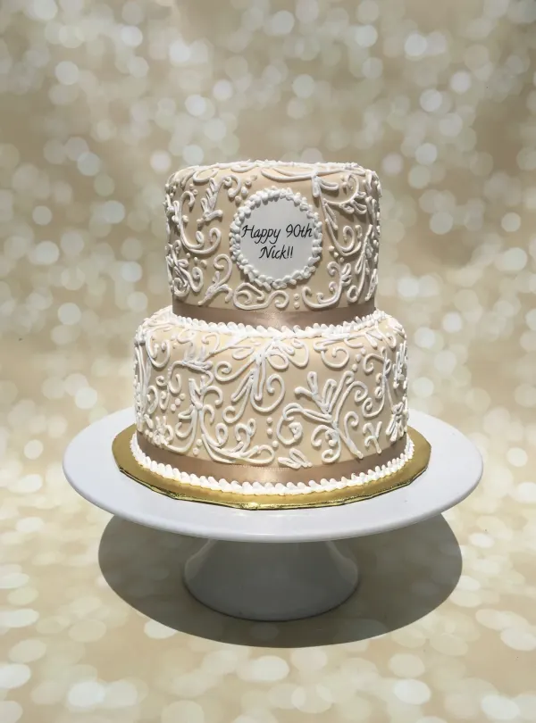 Two-tier champagne cake with white piped scrollwork, satin ribbon and a Happy 90th Nick plaque