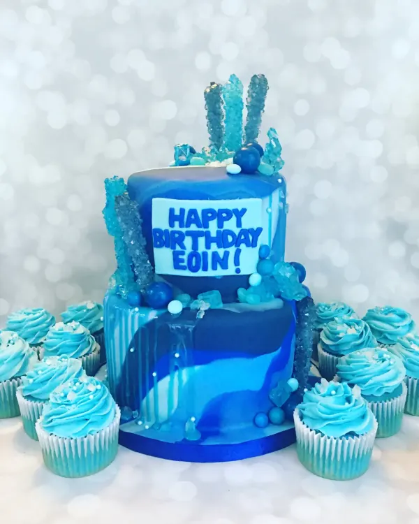 Two-tier blue marbled cake with rock candy sticks and Happy Birthday Eoin, ringed by blue swirl cupcakes