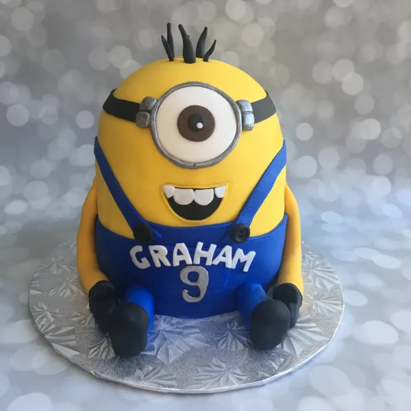 Minion shaped birthday cake with a single goggle eye and blue overalls reading GRAHAM 9