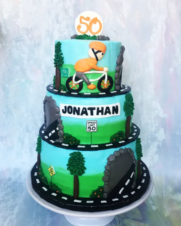 Three-tier cycling cake with roads winding past redwoods, a Speed Limit 50 sign and JONATHAN