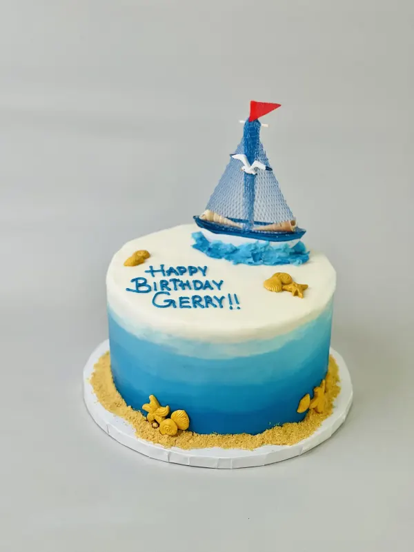 Blue ombre nautical cake with a sailboat topper, gold shells, crumb sand and Happy Birthday Gerry