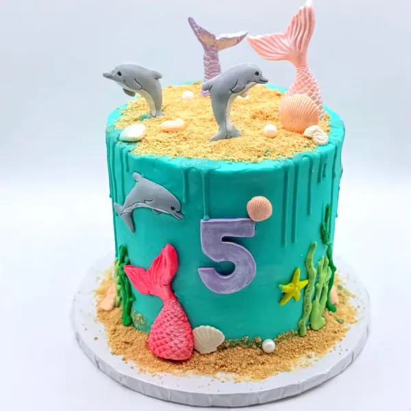 Turquoise cake with dolphin figures, mermaid tails, shells and a sand crumb top with a number five