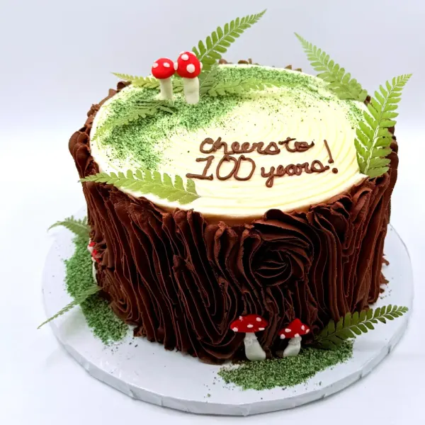 Tree stump cake with piped bark, a mossy top, red mushrooms and ferns, lettered Cheers to 100 years