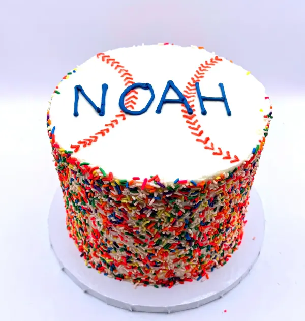 White cake with baseball stitching and Noah lettered on top over sides coated in rainbow sprinkles