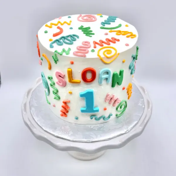 White cake with colorful piped squiggles and doodles, lettered Sloan with a blue number one