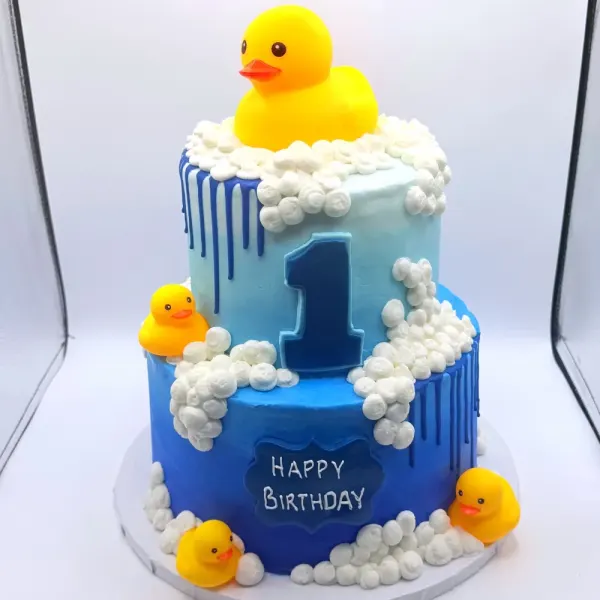 Two tier blue cake with a large rubber duck, fondant bubbles, small ducks and a number one