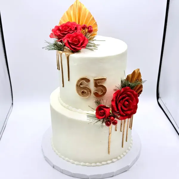 Two tier white cake with gold drips, red roses, a gold palm fan and a gold 65