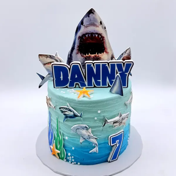 Blue ocean cake with a shark head topper, shark prints around the sides, seaweed and a number seven