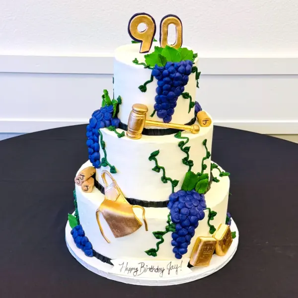 Two tier white cake with purple grape clusters, green vines, gold wine tools and a gold 90 topper