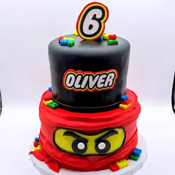 Two tier cake with black and red tiers, brick studs, a ninja mask face and a number six topper