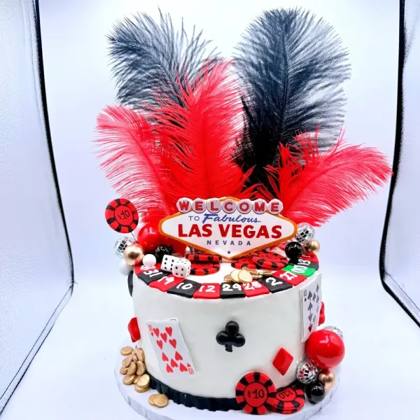 Casino cake with a Las Vegas sign topper, red and black feathers, a roulette wheel, cards and chips