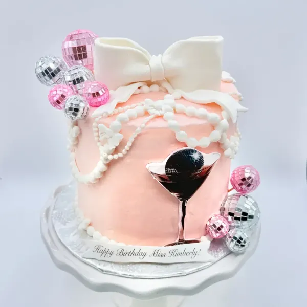 Blush cake with a white fondant bow, pearl strands, disco ball baubles and a martini glass