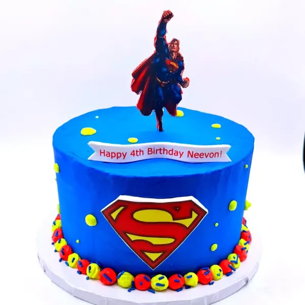 Blue cake with a caped superhero figure, a shield emblem and a colorful bead border