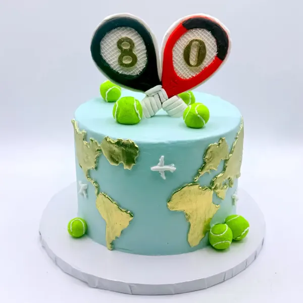Pale blue cake with gold map continents, a small plane and two tennis racket toppers reading 80