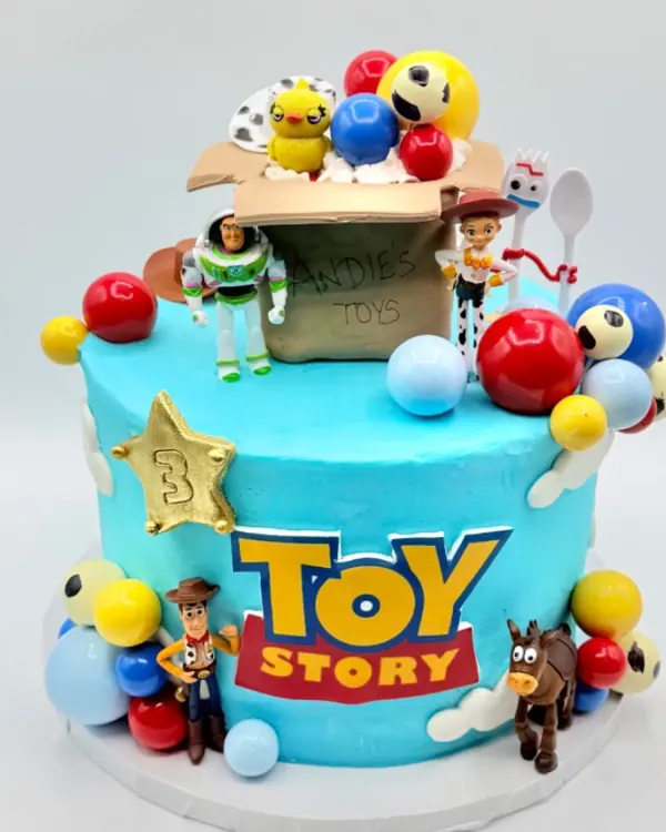 A round cake in sky blue buttercream with a gold sheriff's star numbered three, a hand painted toy story logo, small toy figurines standing around the base and on top, and an open toy box topper filled with colorful balls, photographed against a plain white backdrop.
