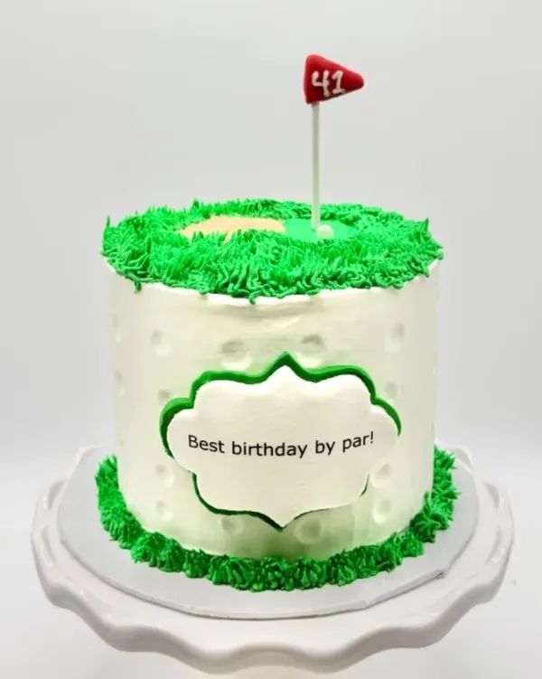A round cake with dimpled white buttercream sides resembling a golf ball, topped with a piped green putting green, a sand trap, and a small red flag reading 41, with a plaque on the front reading Best birthday by par, displayed on a white cake stand.