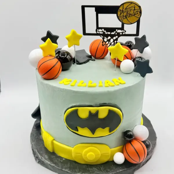 Round sage green cake with a superhero crest and yellow belt design on the front, topped with mini basketballs, a basketball hoop and net topper, and hand shaped stars, with the name Killian piped on top.