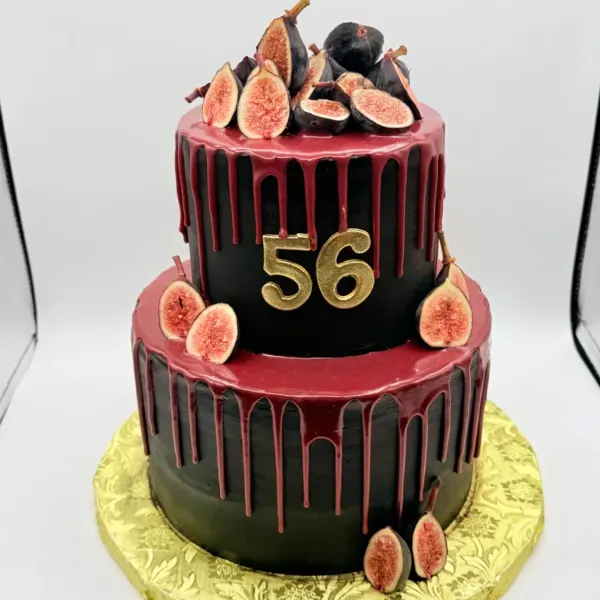 A two tier round cake with black chocolate frosting and a dripping burgundy glaze, topped and surrounded with fresh halved figs and a gold number 56, set on a gold patterned cake board.