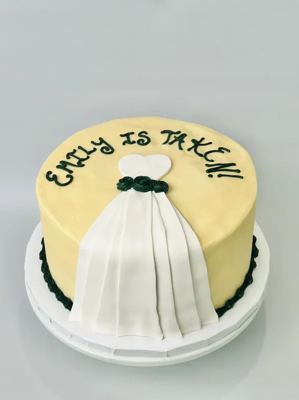 Round ivory cake with a white fondant wedding gown and green lettering reading EMILY IS TAKEN